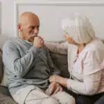 Elderly man looking pensive on a sofa, hand near mouth, as an elderly woman sits beside him, offering comfort with her hand on his shoulder. Calm, supportive mood.