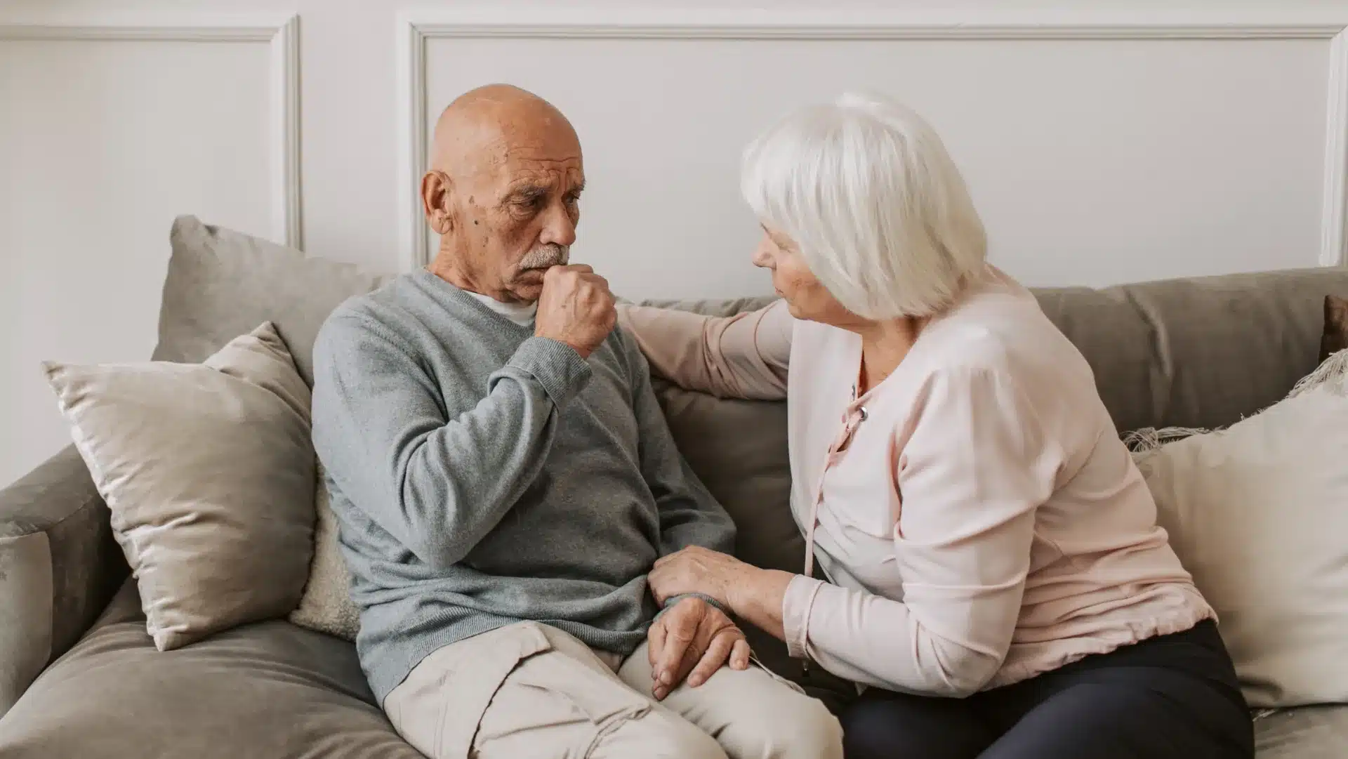 Elderly man looking pensive on a sofa, hand near mouth, as an elderly woman sits beside him, offering comfort with her hand on his shoulder. Calm, supportive mood.