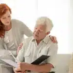 A young woman with red hair comforts an elderly man seated with a photo album. They appear to share a tender moment in a softly lit, cozy room.