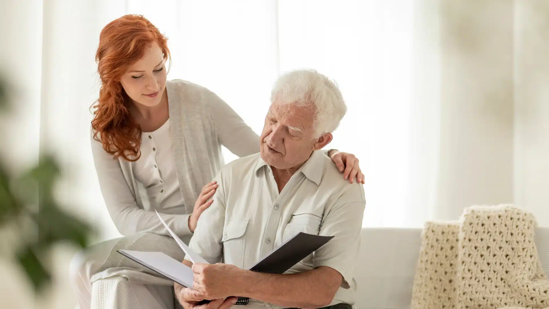 A young woman with red hair comforts an elderly man seated with a photo album. They appear to share a tender moment in a softly lit, cozy room.