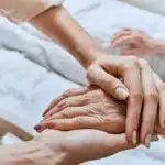 Close-up of a younger person gently holding an elderly person's hand in a comforting manner. Both sit in a hospital bed, conveying care and support.