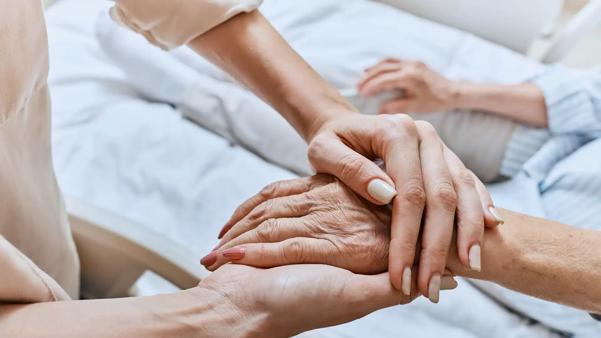 Close-up of a younger person gently holding an elderly person's hand in a comforting manner. Both sit in a hospital bed, conveying care and support.