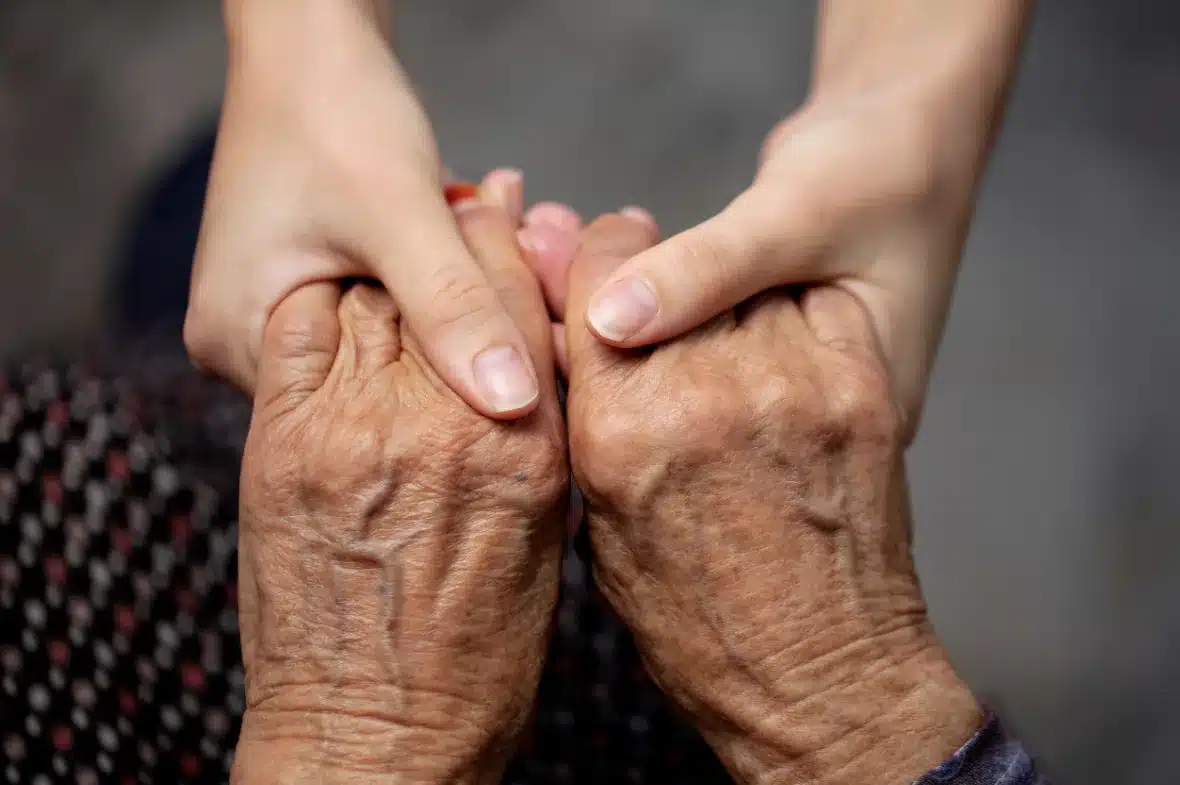 Two hands of a young person gently hold the hands of an elderly person, conveying warmth, care, and connection against a soft, blurred background.