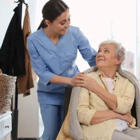 A caregiver in blue scrubs gently places a blanket on an elderly woman in a chair, both smiling warmly, conveying care and companionship in a cozy room.