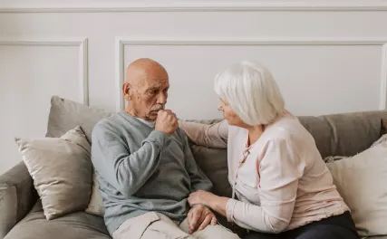 Elderly couple sitting on a sofa; woman in pink comforting a worried man in gray. The scene conveys warmth and support, set in a cozy living room.