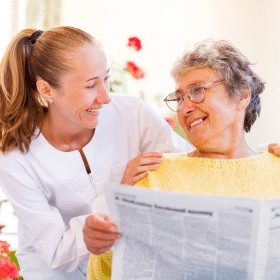 A caregiver smiles at an elderly woman holding a newspaper. They appear happy and engaged. The background is softly lit with red flowers visible.