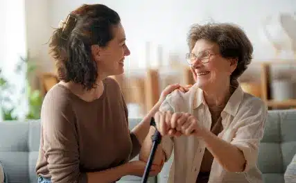 A young woman and an elderly woman with glasses share a joyful moment on a sofa. The elder holds a cane, both smiling warmly at each other in a sunlit room.