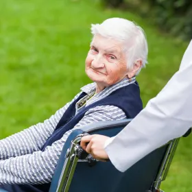 An elderly woman with white hair sits in a wheelchair outdoors on a green lawn, looking content. A person in white pushes the wheelchair.