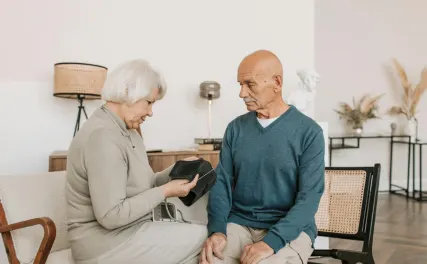 Elderly couple sitting in a bright living room. The woman, holding a black wallet, appears concerned. The man listens intently, creating a thoughtful mood.