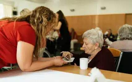 A caregiver assists an elderly woman with a meal at a communal table in a care facility, creating a warm and supportive atmosphere.