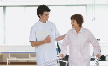 A nurse supports an elderly woman walking with parallel bars in a bright rehabilitation center. Both are smiling, conveying a positive and encouraging atmosphere.