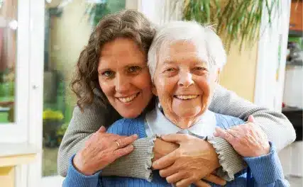 A smiling elderly woman in a blue sweater is lovingly embraced from behind by a younger woman. They stand indoors, conveying warmth and affection.