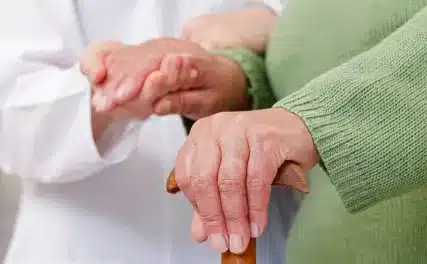 An elderly person in a green sweater holds a cane, while a caregiver in a white coat gently supports their hand, conveying care and support.