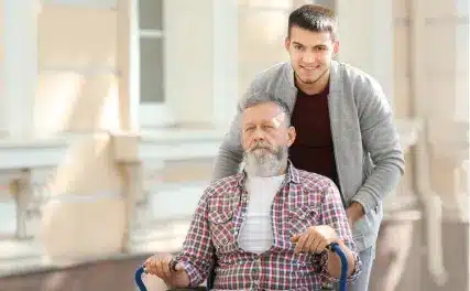 Young man smiles while pushing an elderly man in a wheelchair outside a sunlit building. The elder appears content, wearing a plaid shirt and white tee.
