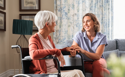 Two women sitting in a cozy living room, smiling warmly at each other. One is elderly, in a wheelchair, while the other sits beside her, holding her hand.