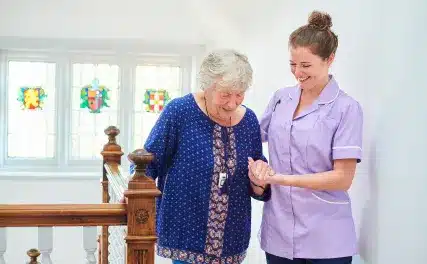 A nurse in a purple uniform smiles while assisting an elderly woman with gray hair in a blue dress down a staircase. Bright, stained glass windows are in the background.