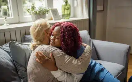 Two people hugging on a gray sofa in a cozy room. One has short blonde hair, the other has curly red hair. The embrace conveys warmth and comfort.