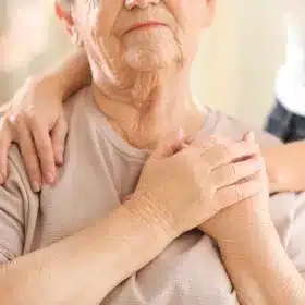 An elderly person with a gentle smile holds their hands over their heart. Another pair of hands rests supportively on their shoulders, conveying warmth and care.