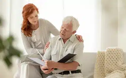 A woman with red hair gently supports an elderly man reading a folder on a sofa. The scene is warm and caring, with soft lighting and cozy decor.