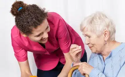 A caregiver in a pink uniform warmly assists an elderly woman with short white hair, offering her a pill. The scene conveys care and support.