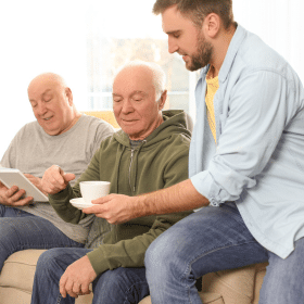 A young man in casual clothes serves coffee to two elderly men sitting on a sofa, one looking at a tablet. The scene conveys warmth and companionship.