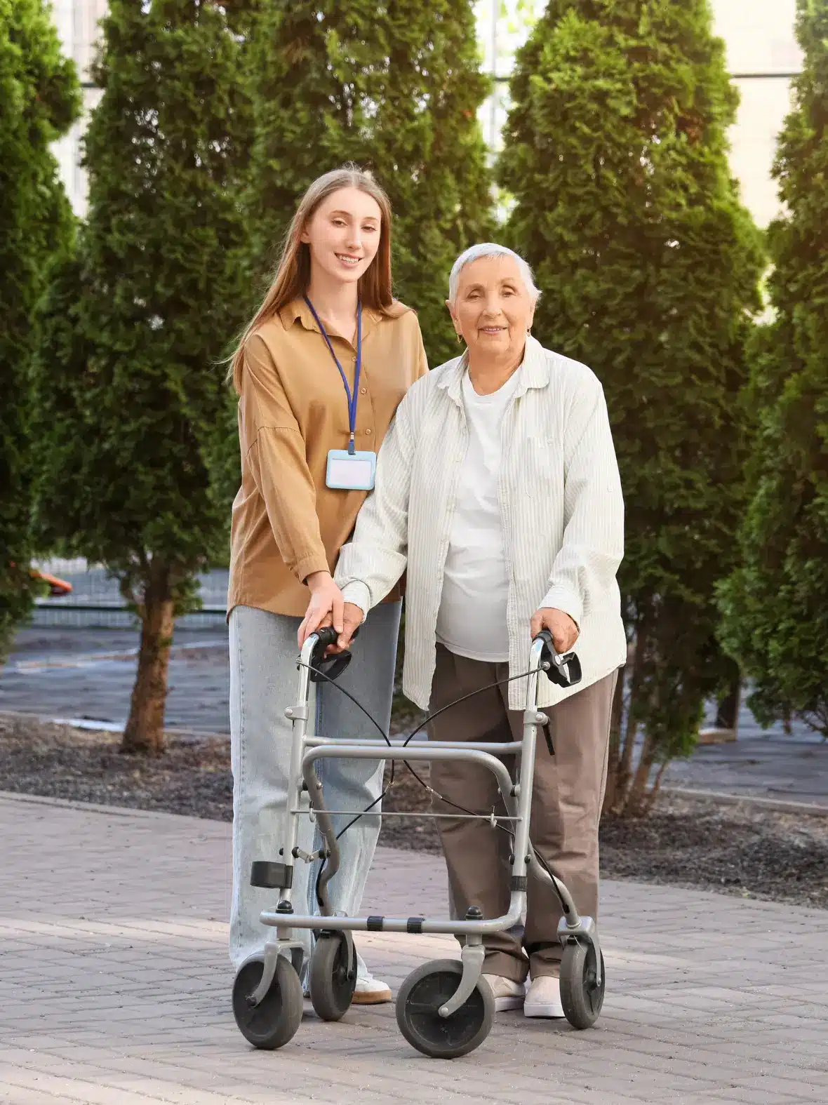 A young woman assists a smiling elderly woman with a walker, set against a backdrop of tall trees. The scene conveys warmth and support.