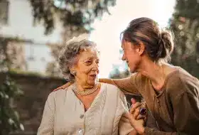 A smiling elderly woman in a pearl necklace and light sweater sits outdoors, warmly interacting with a younger woman in a brown top. Sunlight filters through trees.