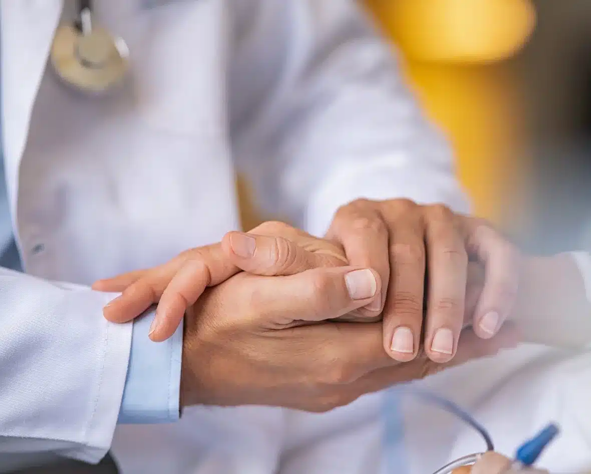 A doctor in a white coat gently holds a patient's hand, conveying comfort and empathy. A stethoscope is visible, highlighting the medical context.