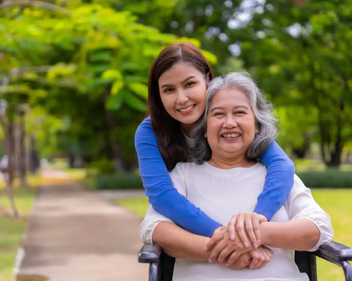 A woman joyfully embraces an elderly woman in a wheelchair in a sunny park. Both smile warmly, conveying love and happiness amid lush green trees.
