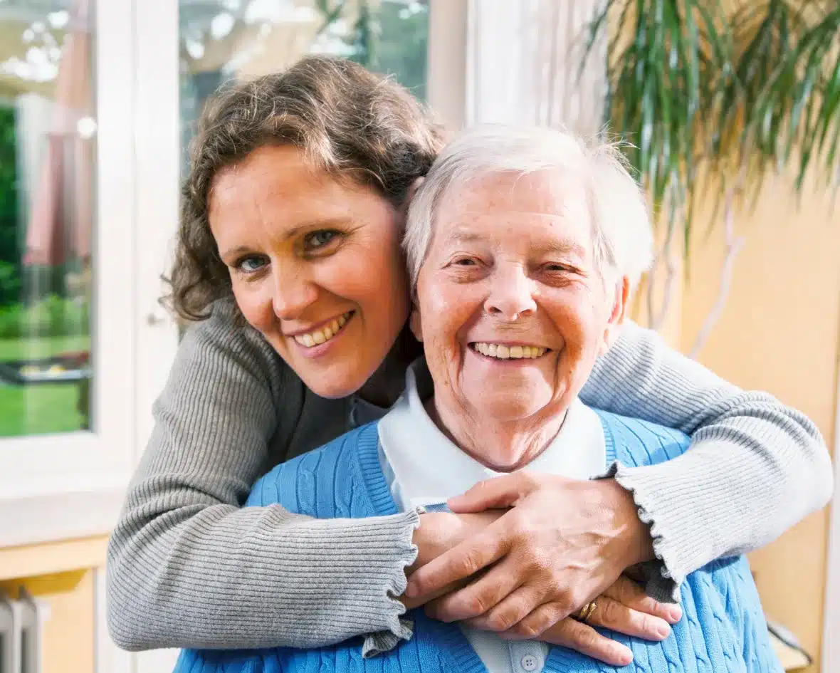 A woman with brown hair hugs an elderly woman with short white hair. Both smile warmly. They are indoors with a window and plant in the background.