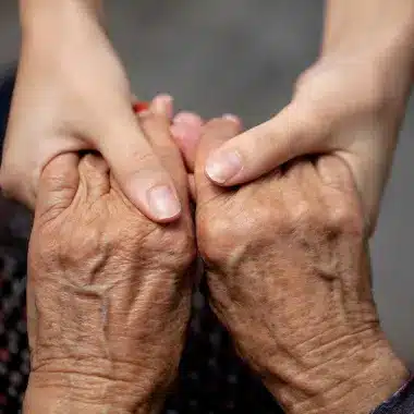 Two pairs of hands: a younger pair gently holding an older pair, conveying warmth and care. The background is soft and neutral, highlighting connection.