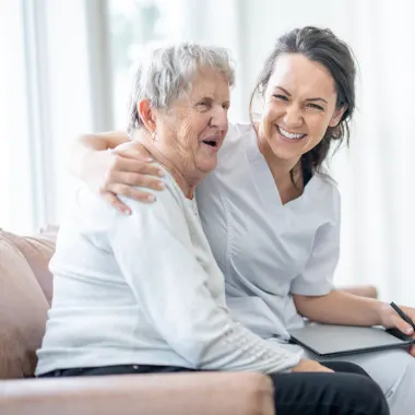 A nurse and an elderly woman sit together on a couch, both smiling warmly. The nurse has her arm around the woman, conveying care and companionship.