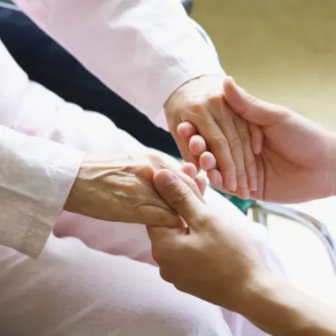 Two hands gently hold an older person's hand, conveying comfort and support. The older person is in a light pink garment, suggesting a caring environment.
