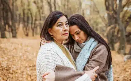 A woman and a younger woman embrace in a forest with autumn leaves, conveying support and comfort. The atmosphere is calm and reflective.