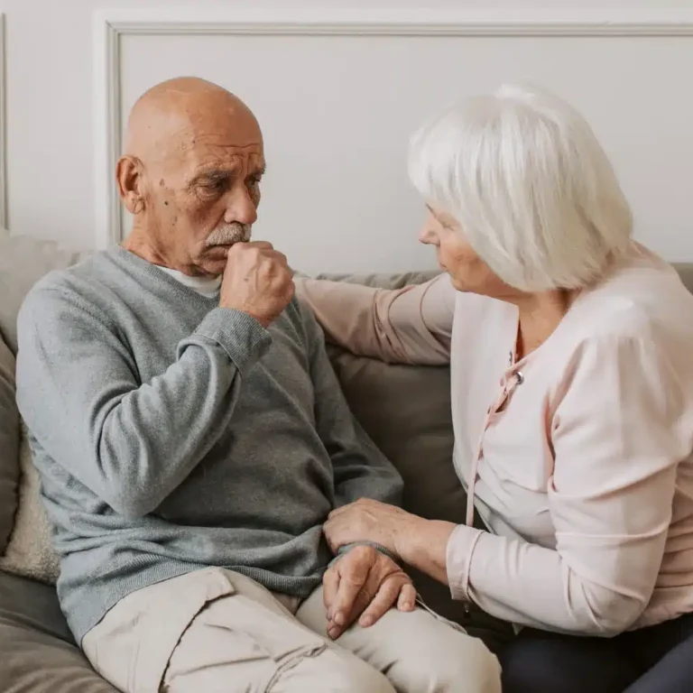 Elderly man looking pensive on a sofa, hand near mouth, as an elderly woman sits beside him, offering comfort with her hand on his shoulder. Calm, supportive mood.