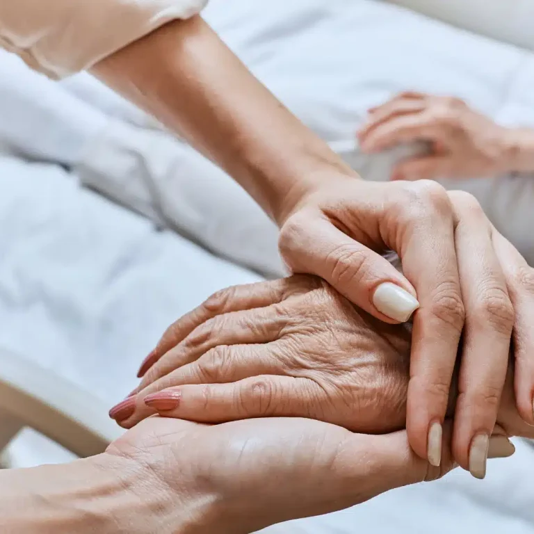 Close-up of a younger person gently holding an elderly person's hand in a comforting manner. Both sit in a hospital bed, conveying care and support.